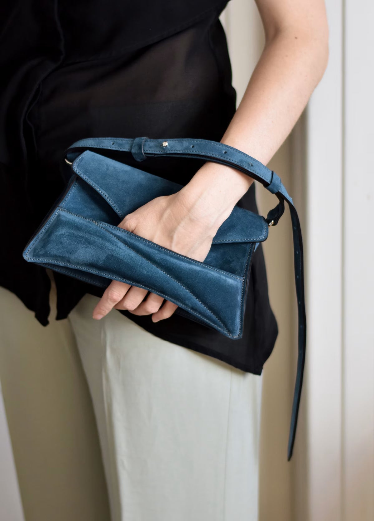 Lindasieto sculptural blue suede calf leather Link Bag in powdery, muted azure blue, designer shoulder bag worn in hand with minimal black outfit. Magyar kortárs designer kék velúr bőr alkalmi clutch táska.