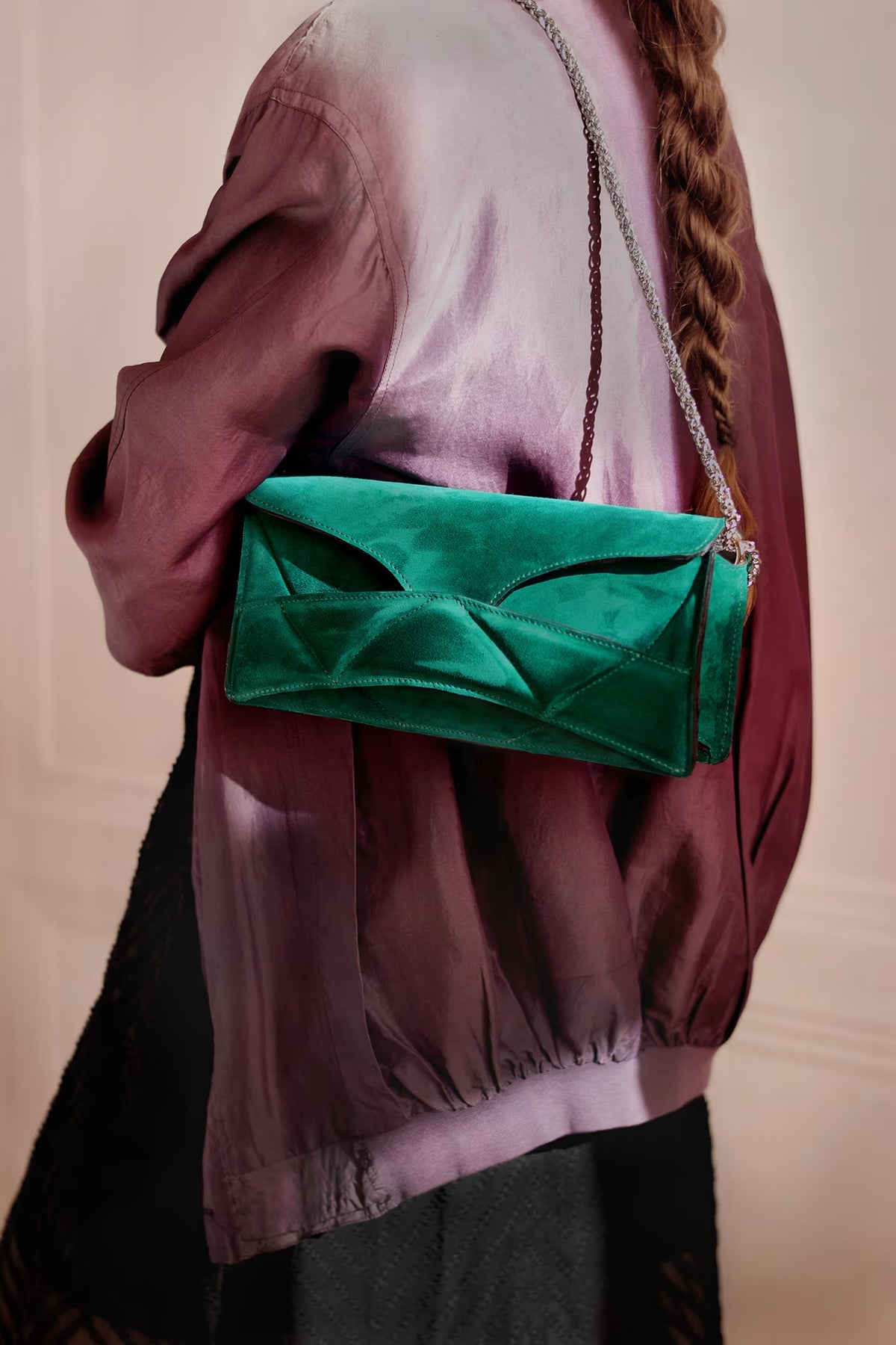 Person in mauve silk jacket and black skirt wearing LINDASIETO's Abstract Heart Bag on the shoulder, in emerald green suede leather, sculptural clutch bag with layered heart pattern, with a silver chain strap.