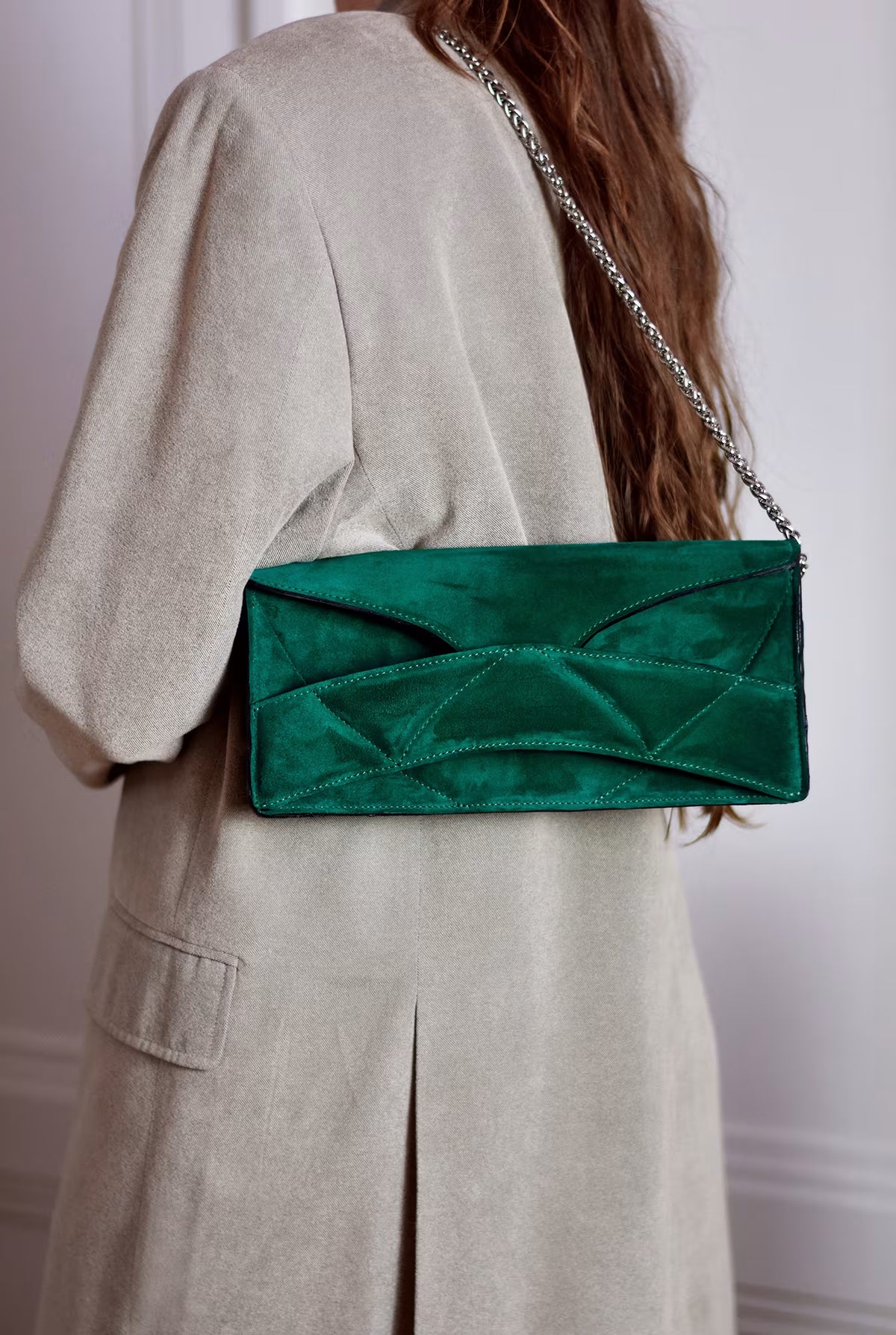 Person in beige jacket wearing LINDASIETO's Abstract Heart Bag on the shoulder,  designer clutch bag with layered heart pattern, sculptural wrist strap with a silver chain shoulder strap. in emerald green suede leather.