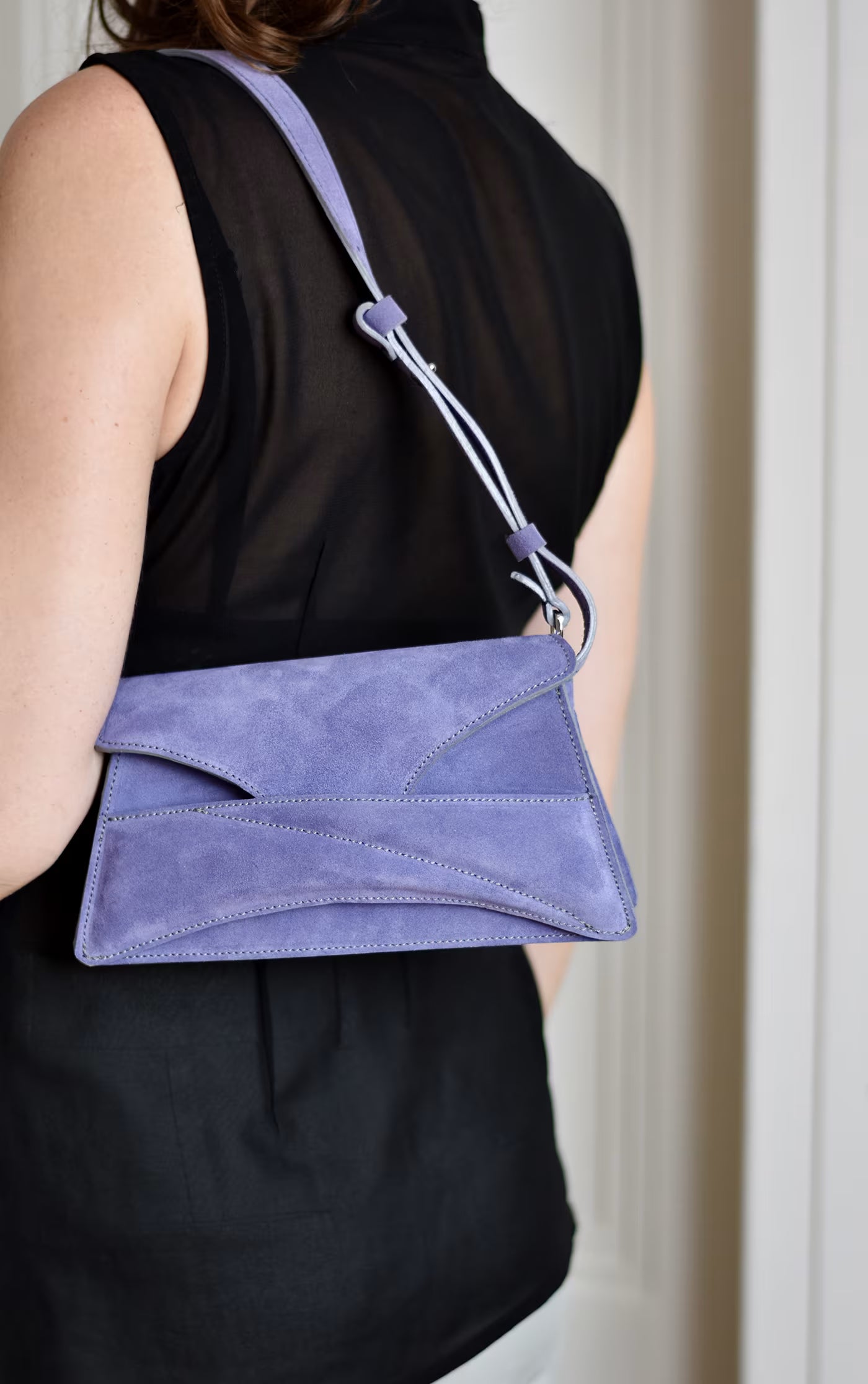 Lindasieto sculptural amethyst lilac suede leather convertible Link Bag, designer handbag with unique wrist strap detail, worn on the shoulder with black elegant minimal outfit. Magyar kortárs designer lila velúr bőr alkalmi clutch táska, vállon viselve, elegáns fekete neutrális öltözékkel