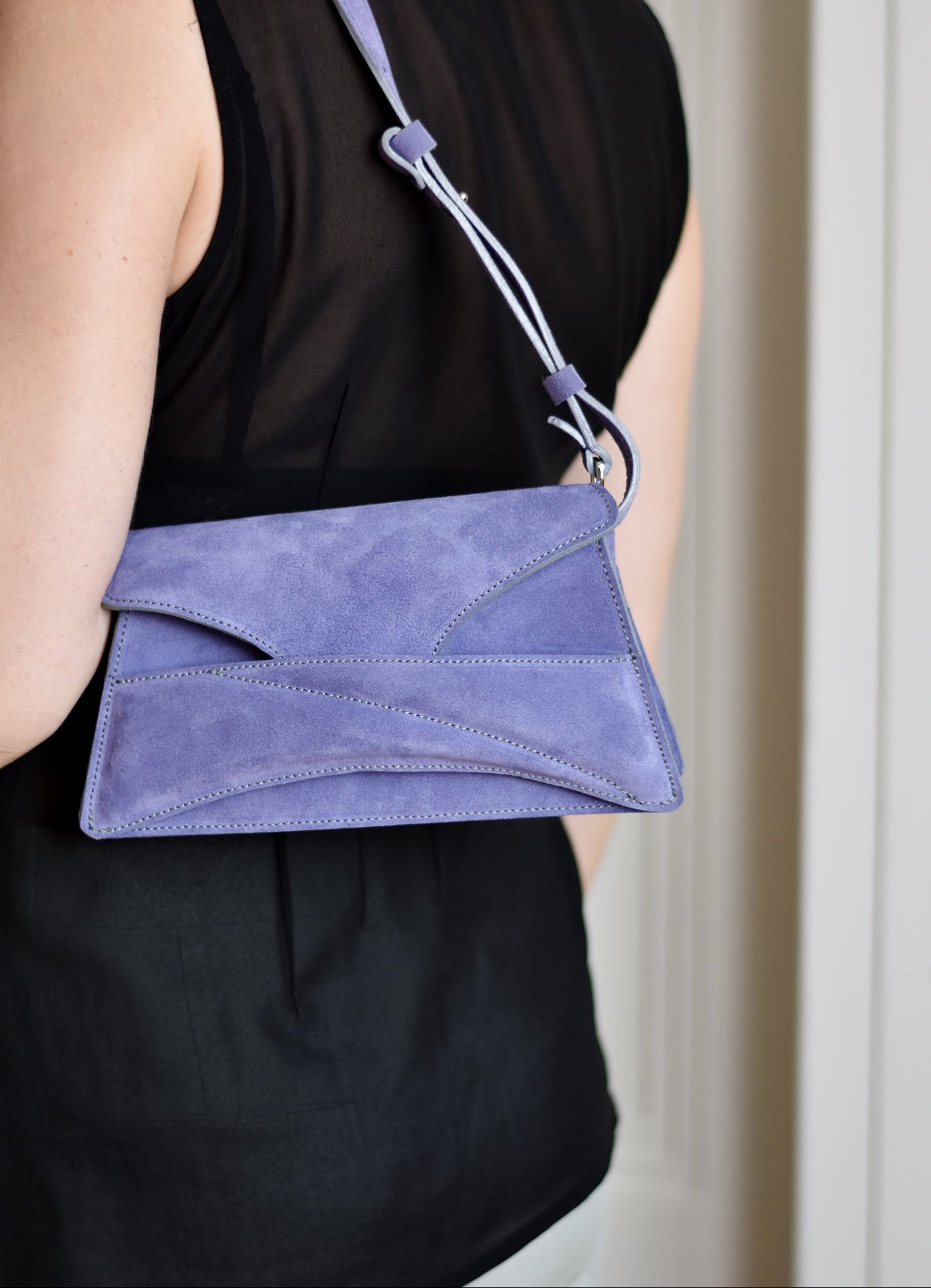 Lindasieto sculptural amethyst lilac suede leather Link Bag, designer crossbody handbag worn on the shoulder with black elegant minimal outfit. Magyar kortárs designer lila velúr bőr alkalmi clutch táska, kézben viselve, elegáns fekete neutrális öltözékkel