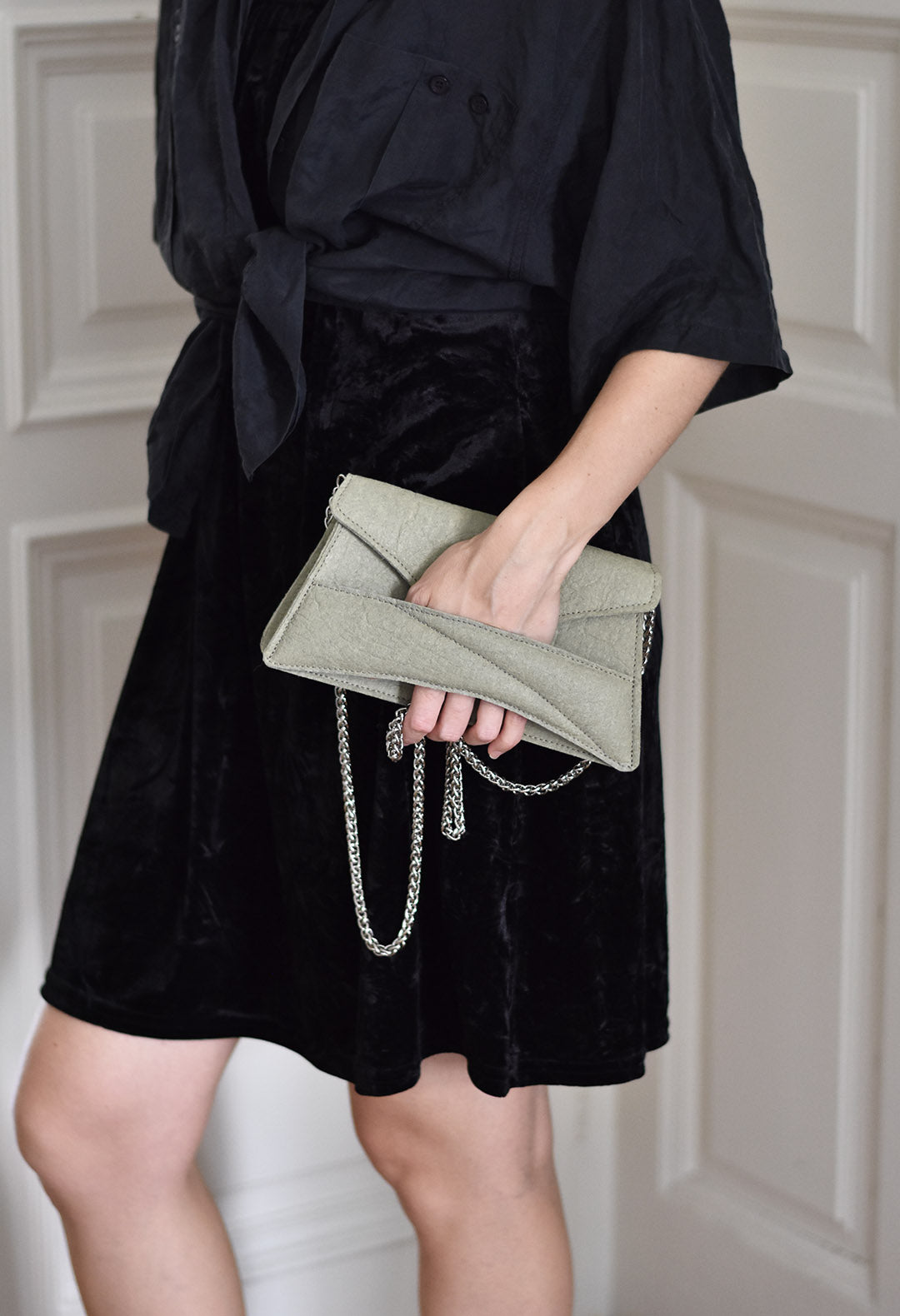 A person wearing a black dress and black shoes, wearing  a vegan Link Bag by LINDASIETO in light, plant-based green, with a chain strap, and sculptural wrist strap in hand.