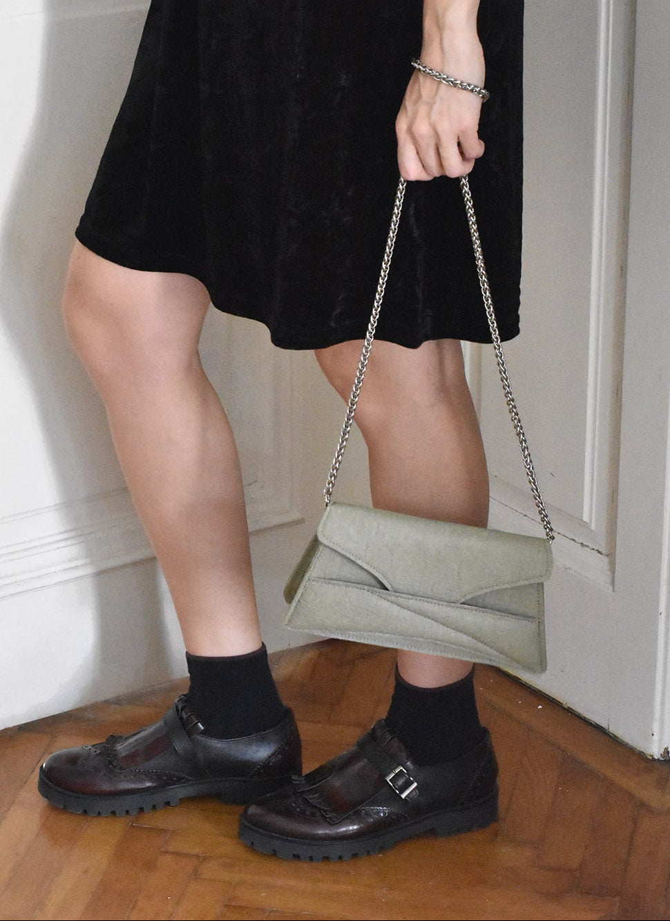 A person wearing a black dress and black shoes, carrying a vegan Link Bag by LINDASIETO in light, plant-based green, with a chain strap.
