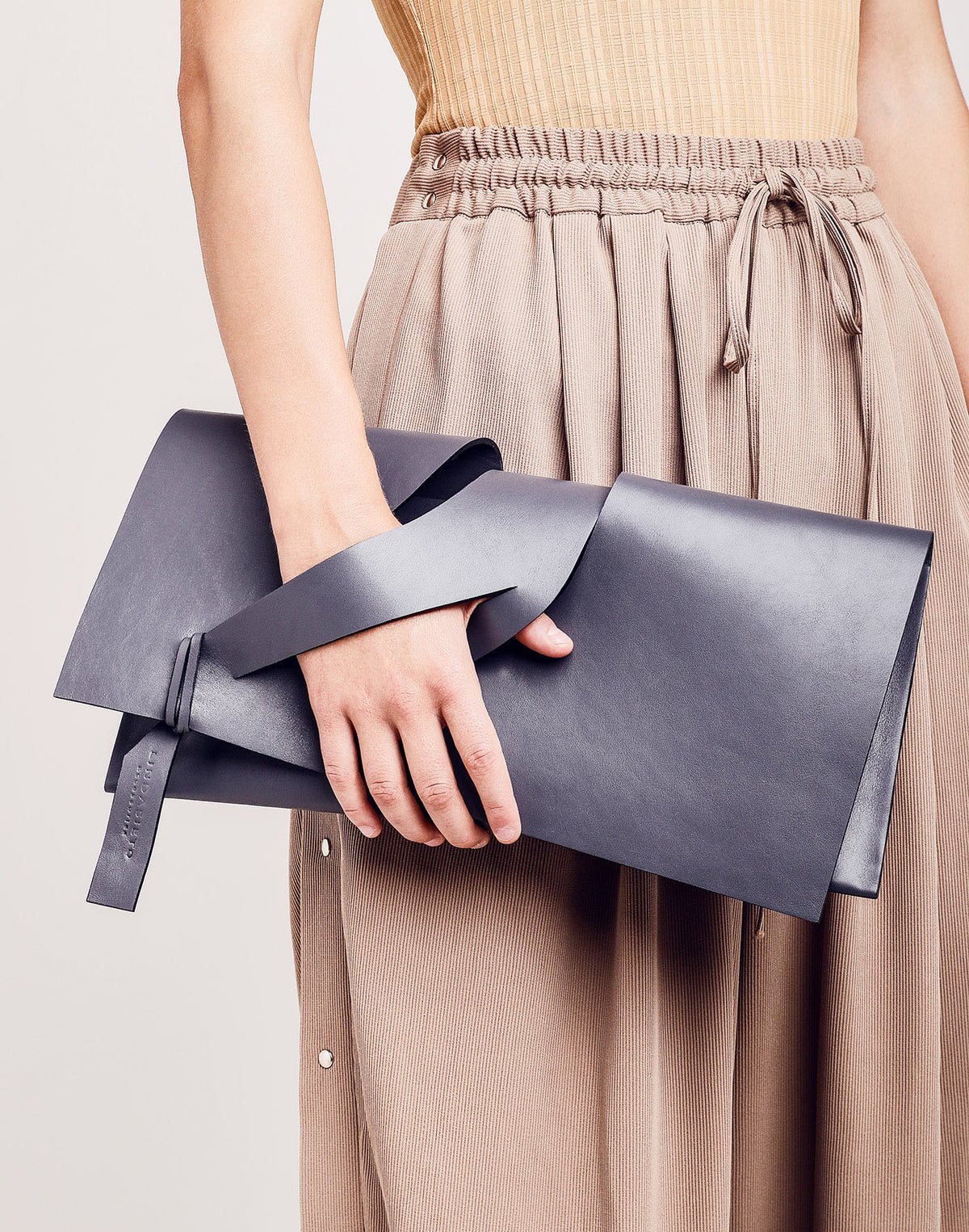 LINDASIETO diagonal extra grey leather architectural clutch bag worn in hand with unique, slit wrist strap design, against timeless, beige trousers .