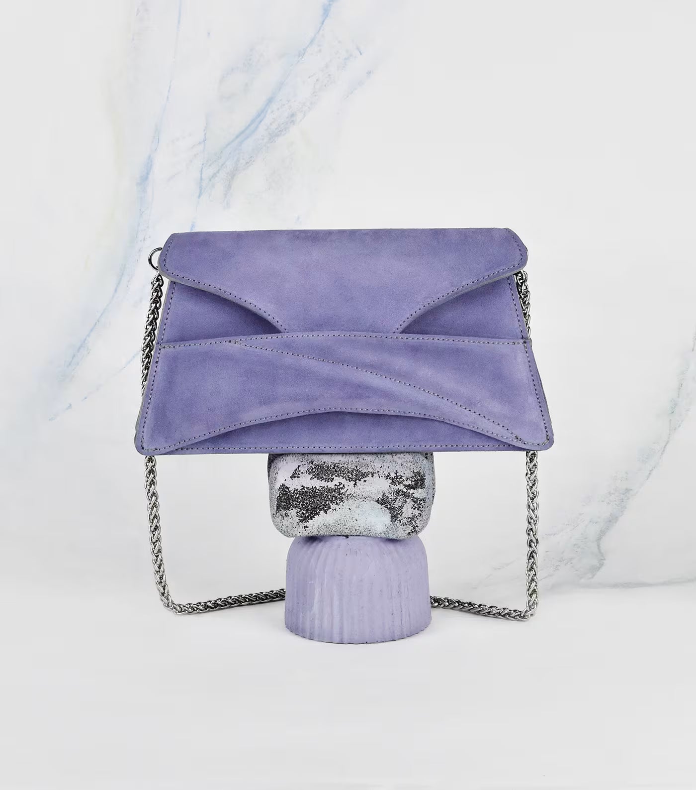 Lindasieto Link Bag, amethyst lilac purple envelope-style clutch with a chain strap, displayed on a decorative stand against a white marble-patterned background. Magyar kortárs designer lila velúr bőr alkalmi női clutch táska