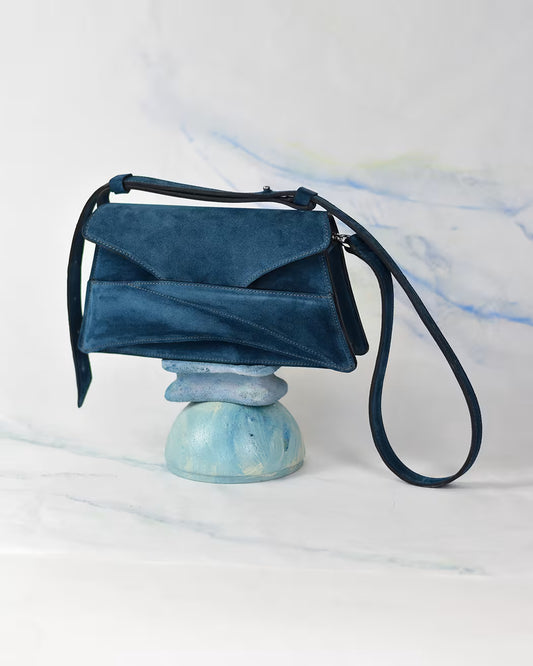 Lindasieto sculptural blue suede leather Link Bag in midnight azure blue, designer handbag on a painted stone pedestal with a blue-green-white pastel artwork background. Magyar kortárs designer kék velúr bőr alkalmi clutch táska