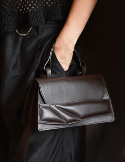 Lindasieto dark brown Mini Balance leather handbag with a sculpted wrist strap and geometric stitched details, carried by a person wearing a minimal, black knit top and elegant black trousers. Magyar designer elegáns csokoládébarna női, uniszex bőr táska.