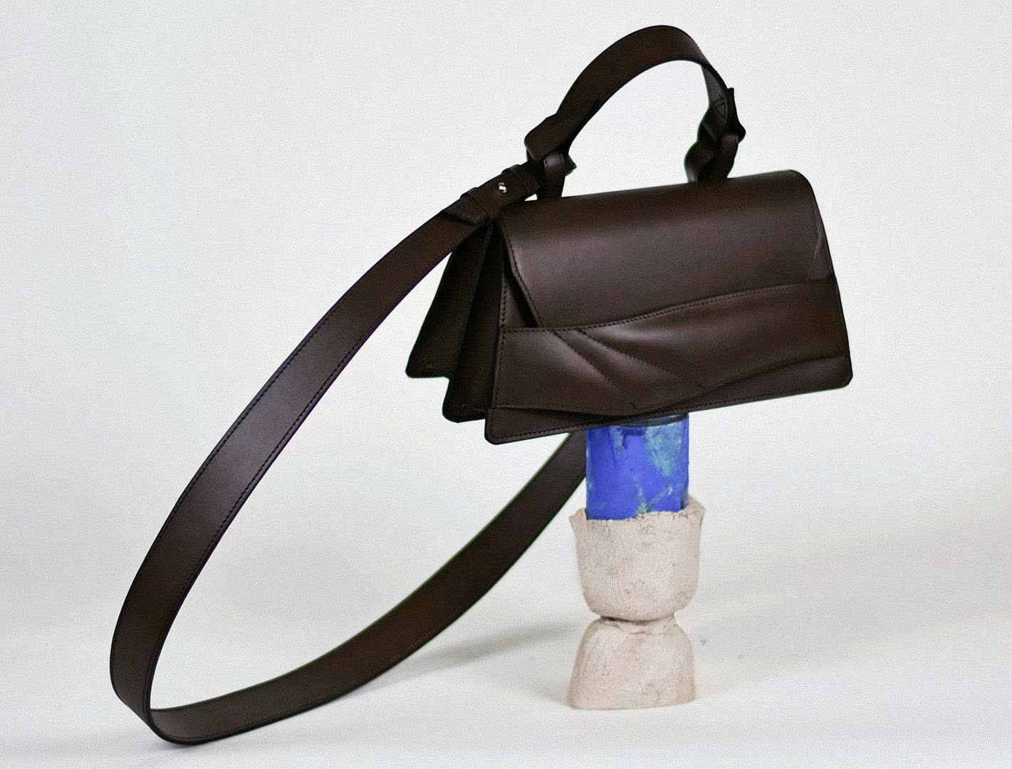 Linda Sieto – Sculptural, quiet luxury leather handbags, mini balance bag in dark brown Italian nappa, placed on ultramarine blue object. magyar designer csokoládé barna bőr contemporary női táska